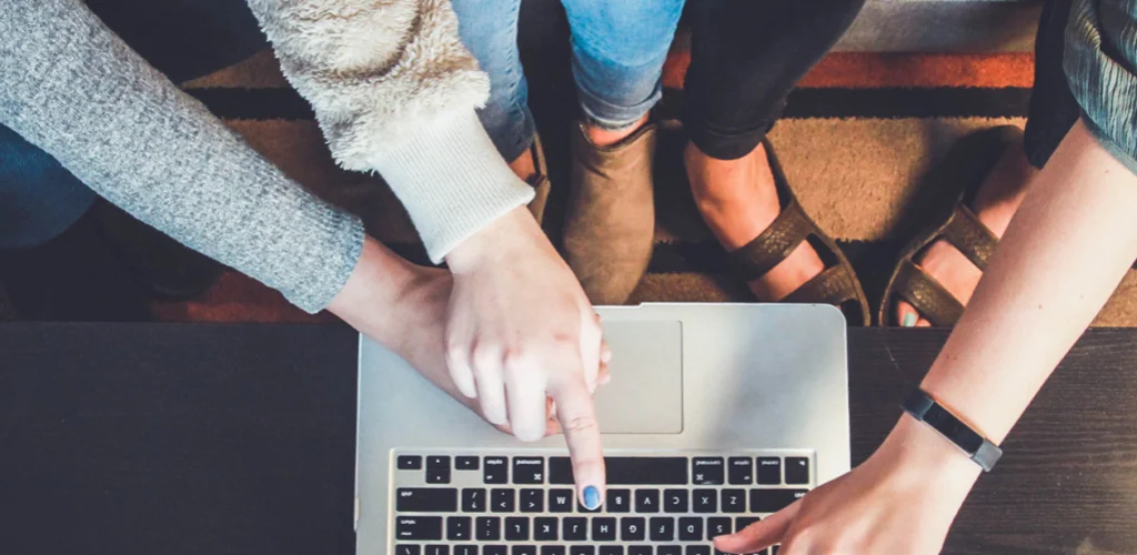 People around a laptop Close-up from above showing several hands, including one wearing a fitness tracker, pointing at a laptop screen, indicating group interaction and problem-solving. This visual captures the essence of how Anchored Virtual Assisting functions as a Strategic Creative & Brand Partner, offering dynamic virtual assistant support and innovative solutions through teamwork.
